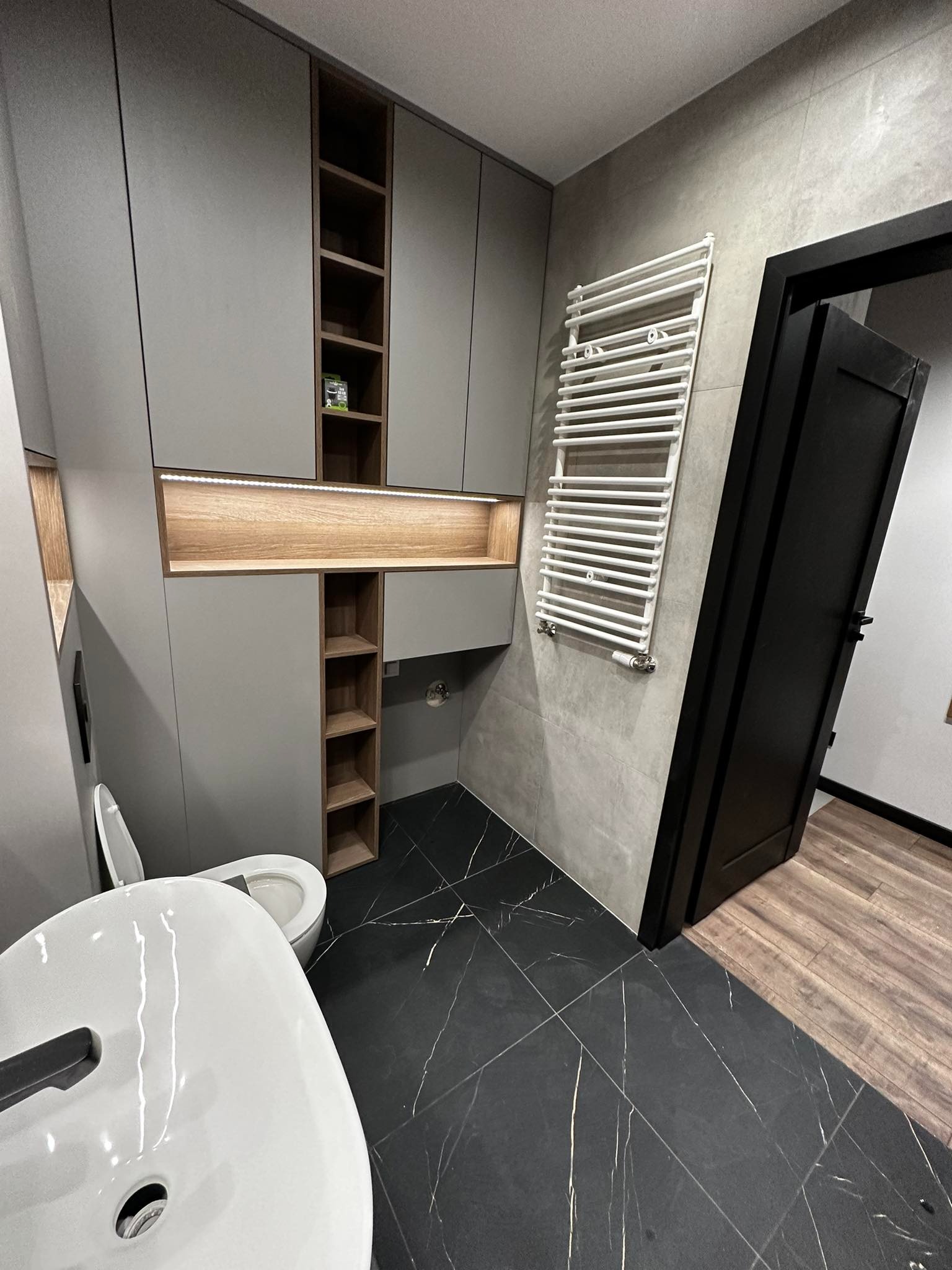 Nowoczesna łazienka z szarą zabudową meblową, podświetlanymi półkami, czarną marmurową podłogą i białym grzejnikiem drabinkowym, widok na otwarte czarne drzwi.