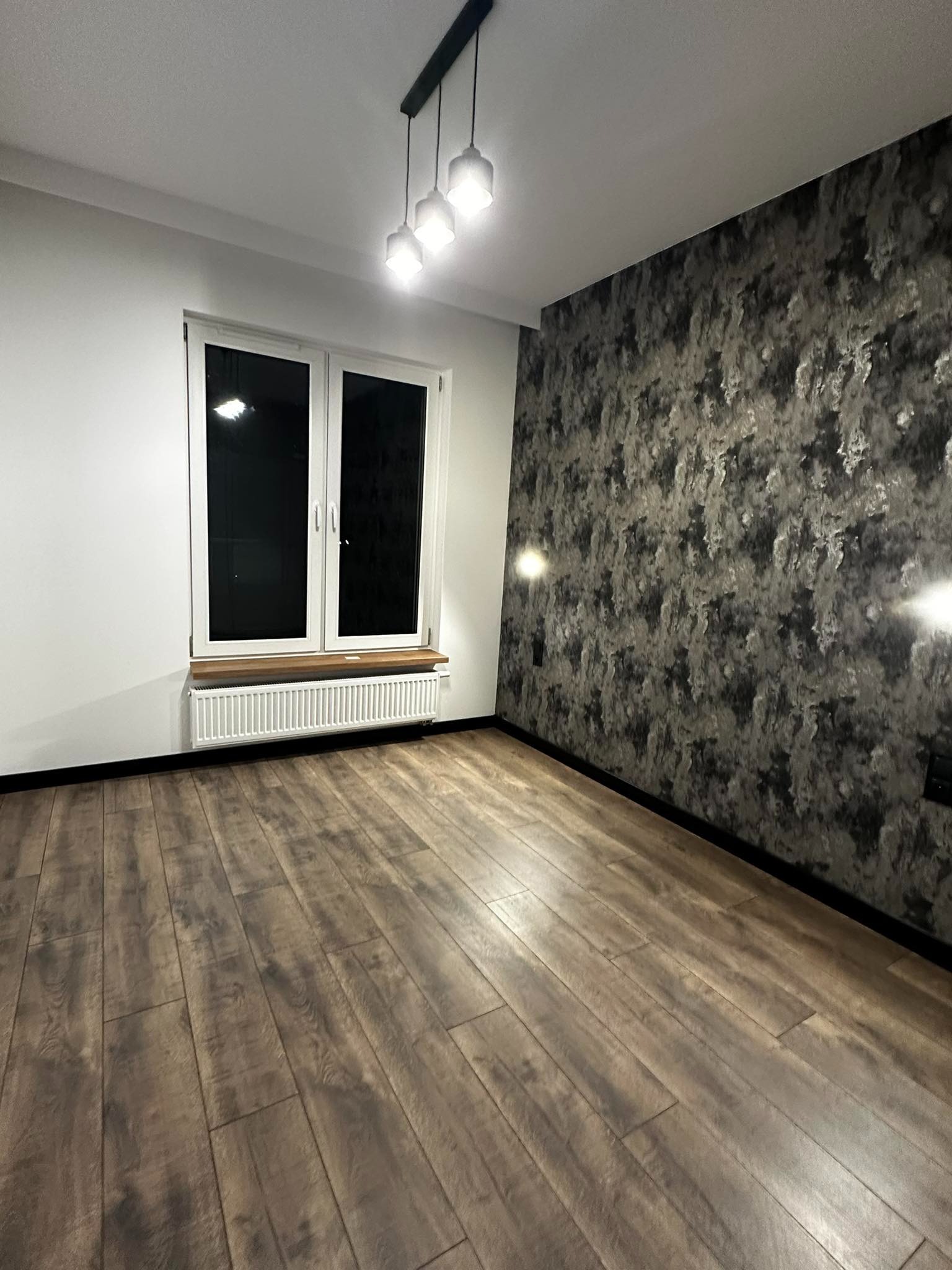 Pusty pokój po remoncie z drewnianą podłogą, grzejnikiem pod oknem i ścianą pokrytą tapetą o teksturze przypominającej kamień.