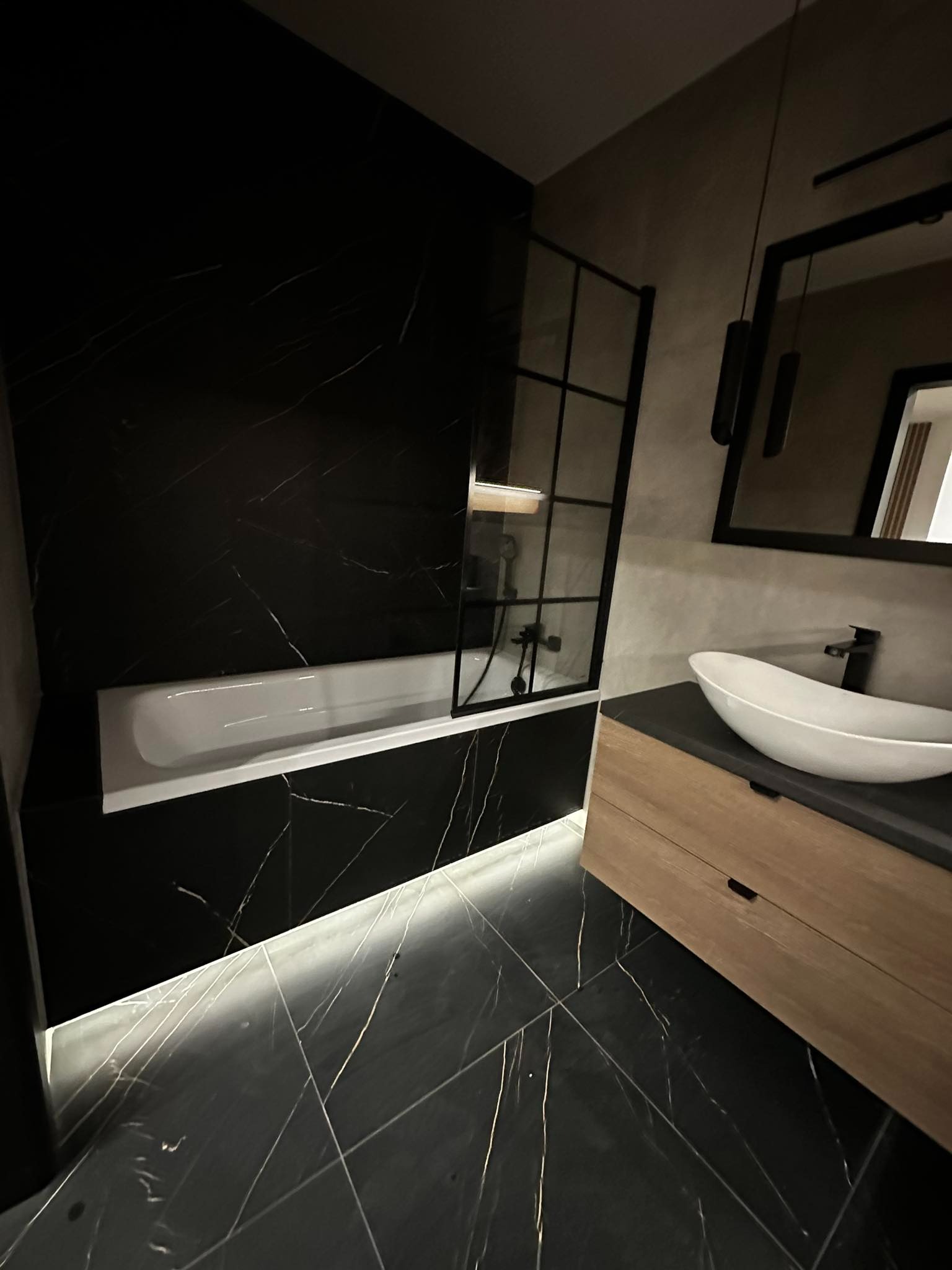 Nowoczesna łazienka w czarno-białej kolorystyce z akcentami drewna: czarne marmurowe płytki z białymi żyłami, biała wanna z parawanem, umywalka nablatowa na drewnianej szafce, oświetlenie LED...