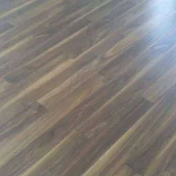 Ukośnie ułożony, ciemny parkiet z wyraźnym usłojeniem, widoczny fragment białej ściany i futryny okiennej.