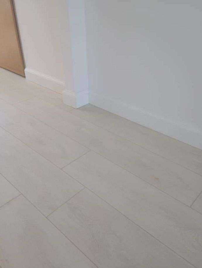 Jasne panele podłogowe ułożone wzdłuż ściany z białą listwą przypodłogową, widok z niskiej perspektywy.