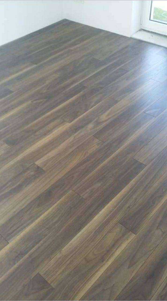 Ukośnie ułożony, ciemny parkiet z wyraźnym usłojeniem, widoczny fragment białej ściany i futryny okiennej.