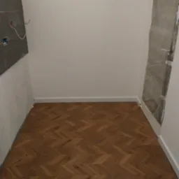 Pok&oacute;j po remoncie: parkiet ułożony w jodełkę, białe listwy przypodłogowe, fragment ściany z instalacją elektryczną i zabezpieczone folią drzwi.