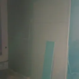 Świeżo pomalowana, gładka ściana w odcieniach szarości, z widocznymi śladami po taśmie malarskiej i niedoskonałościami w dolnej części, oświetlona z prawej strony, z fragmentem listwy...