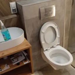 Nowoczesna łazienka z podwieszaną toaletą, drewnianą szafką pod umywalką i marmurowymi płytkami ściennymi, widoczna butelka płynu do czyszczenia i akcesoria na półce.