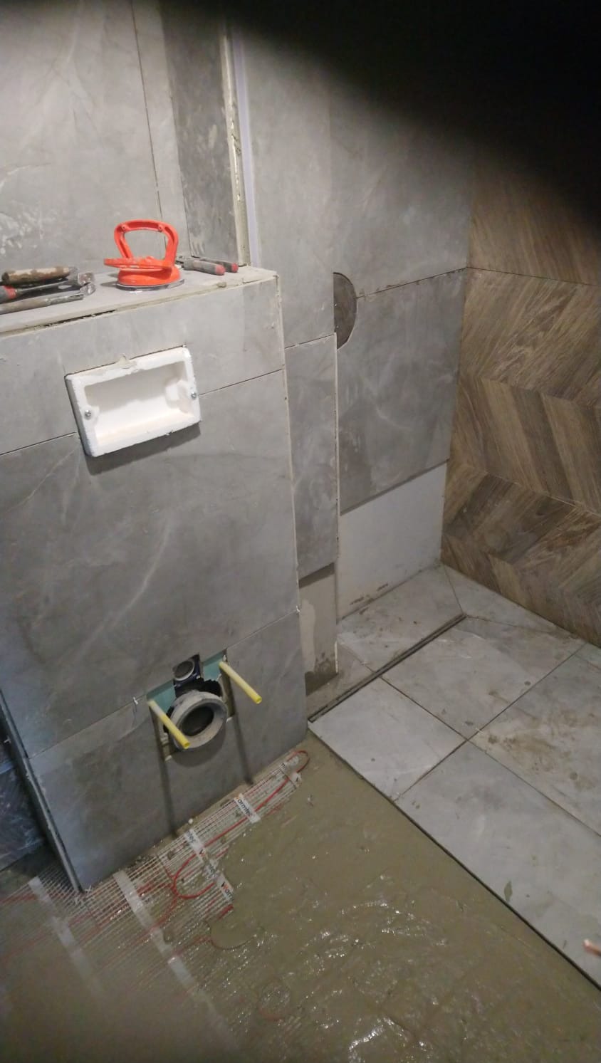 Fragment łazienki w trakcie układania płytek: widoczna instalacja podtynkowego WC, mata grzewcza zalana wylewką, narzędzia do płytek na obudowie stelaża.