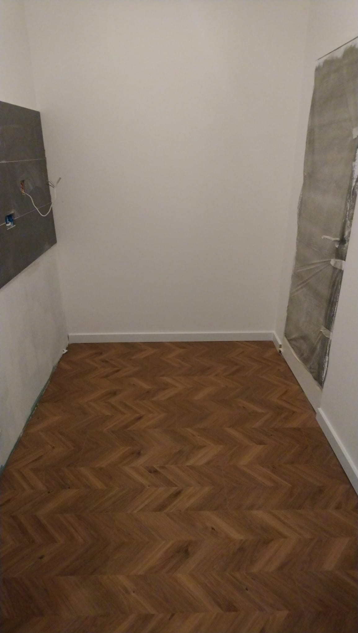 Pokój po remoncie: parkiet ułożony w jodełkę, białe listwy przypodłogowe, fragment ściany z instalacją elektryczną i zabezpieczone folią drzwi.