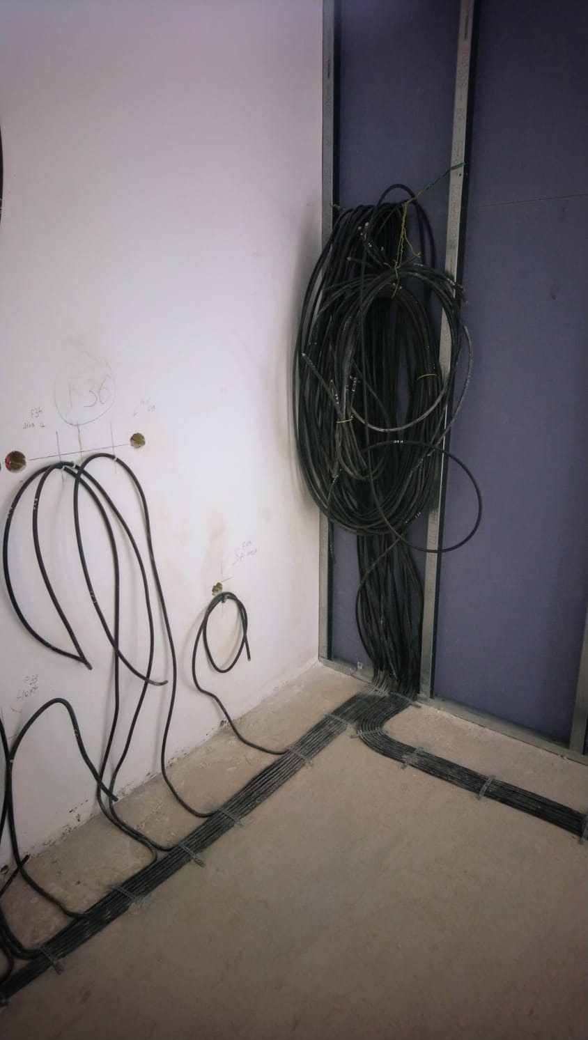 Wiązki kabli elektrycznych prowadzone wzdłuż ściany i podłogi w surowym wnętrzu, widoczne otwory w ścianie z wyprowadzonymi przewodami, ściana z metalowym stelażem.