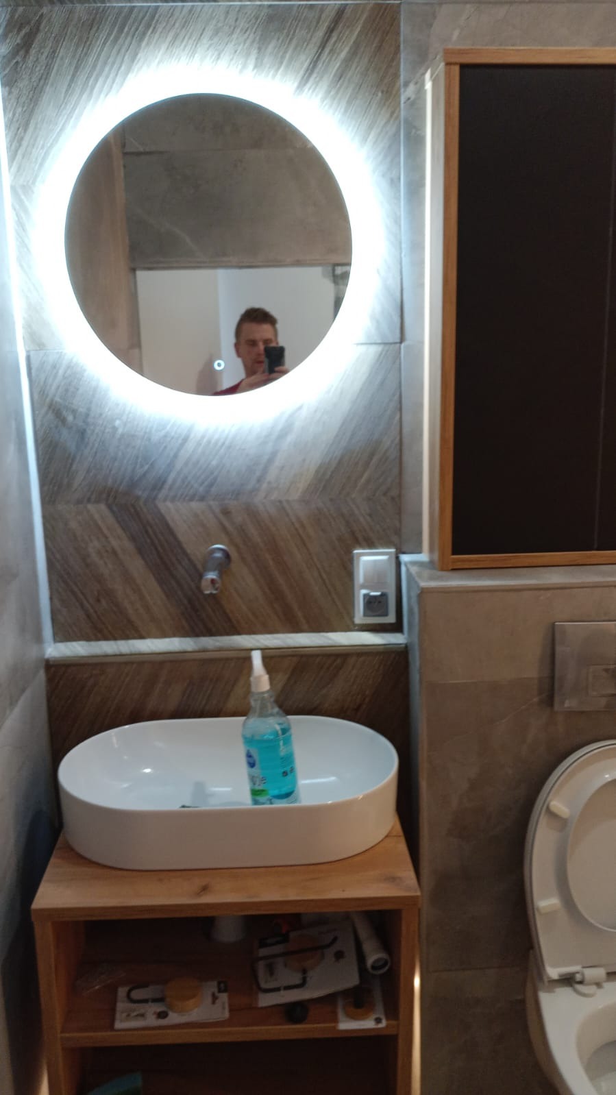 Nowoczesna łazienka z okrągłym lustrem podświetlanym LED, umywalką nablatową i drewnianą szafką. Na blacie umywalki stoi butelka z płynem. W lustrze odbija się osoba robiąca zdjęcie.