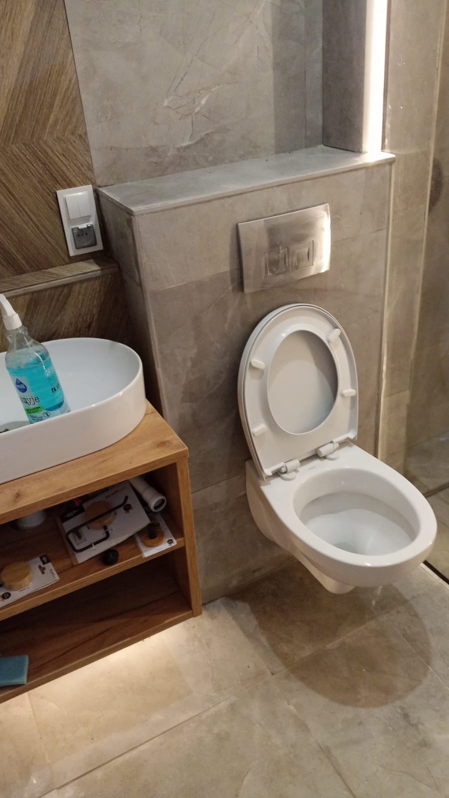 Nowoczesna łazienka z podwieszaną toaletą, drewnianą szafką pod umywalką i marmurowymi płytkami ściennymi, widoczna butelka płynu do czyszczenia i akcesoria na półce.