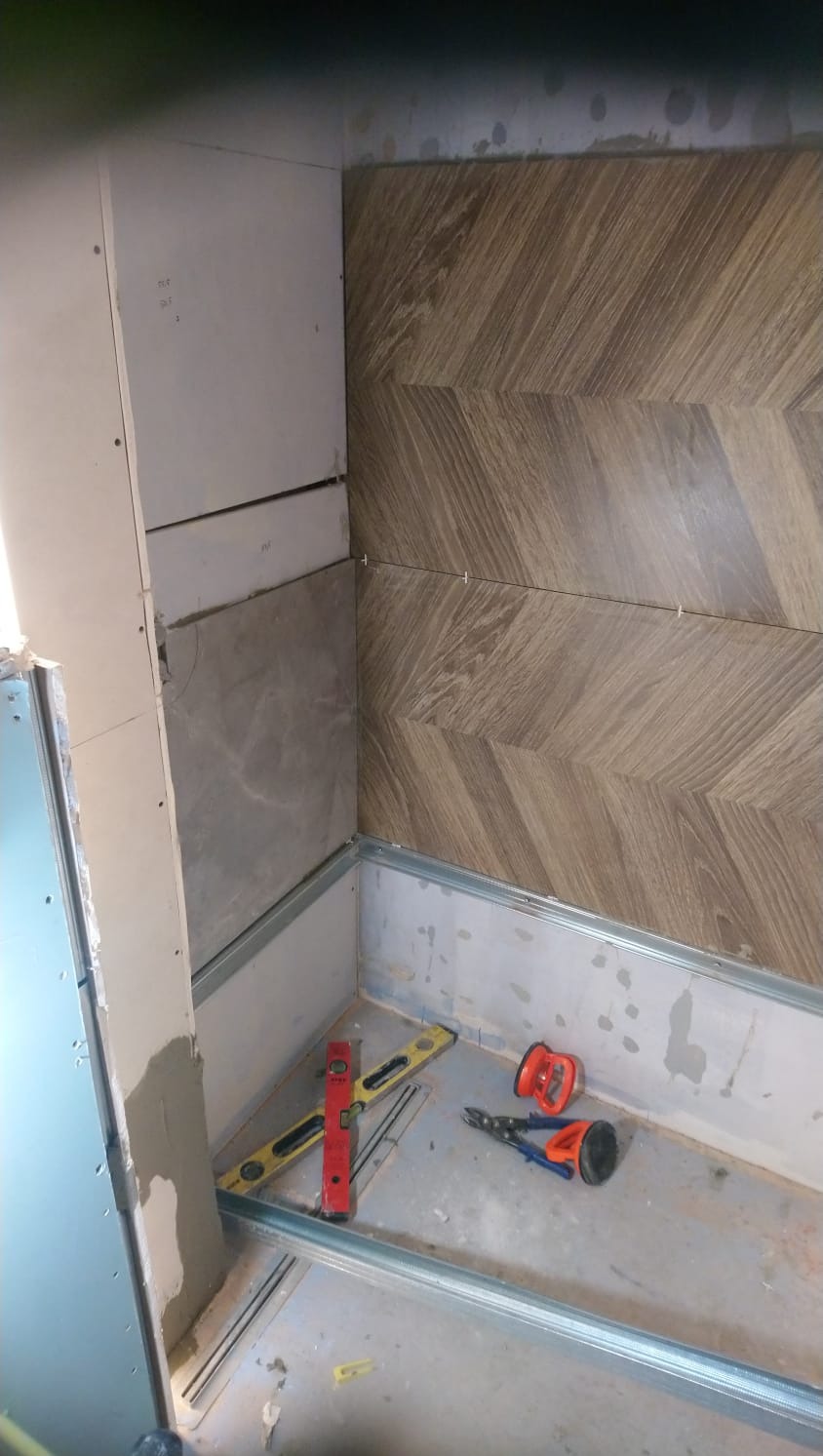 Montaż płytek imitujących drewno w jodełkę na ścianie w trakcie prac, widoczne narzędzia: poziomica, przyssawka do płytek i kombinerki.