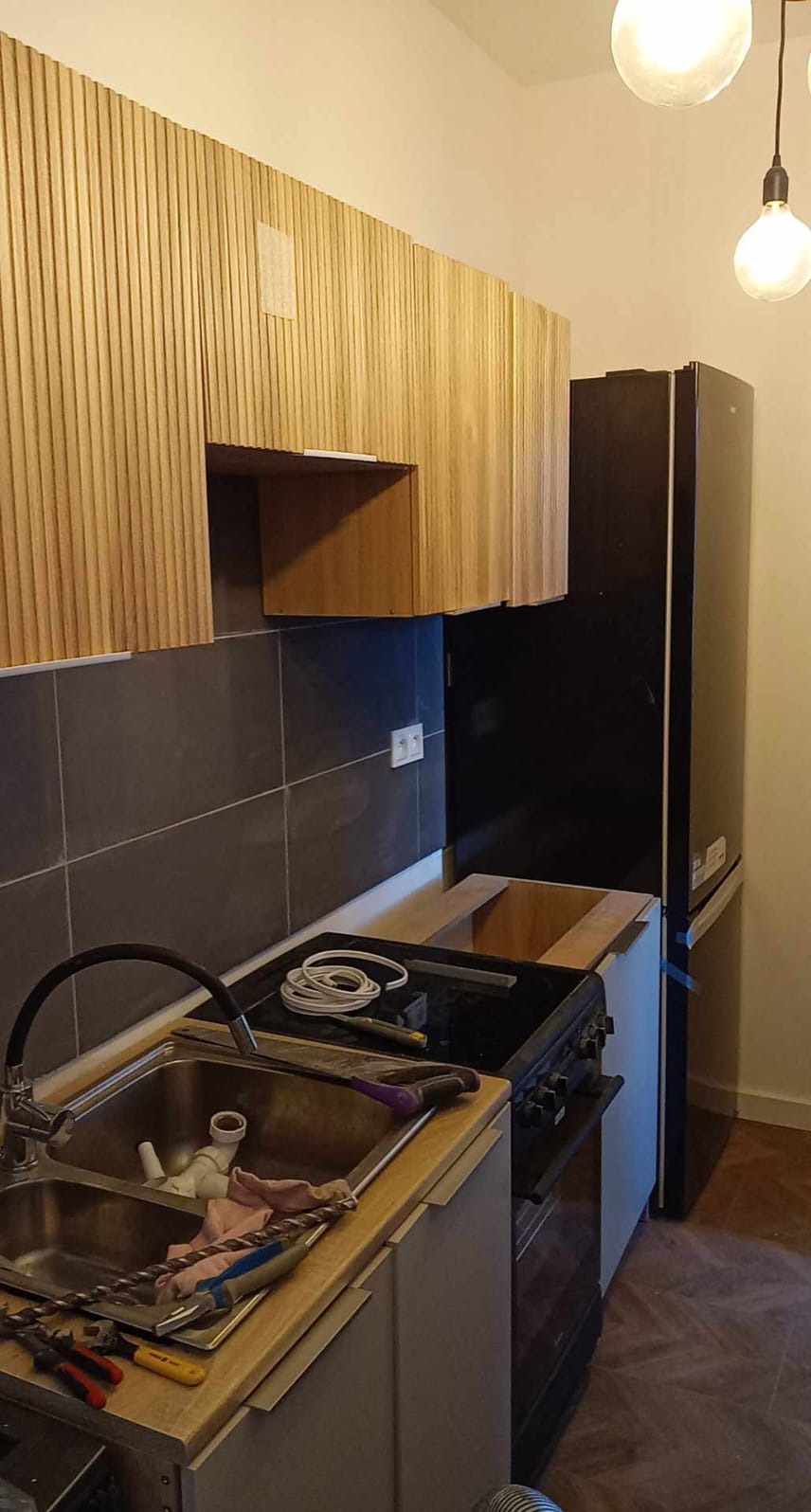 Wnętrze kuchni w trakcie prac: drewniane panele na ścianie, szare płytki, zlewozmywak z narzędziami, płyta grzewcza, lodówka i wiszące lampy.