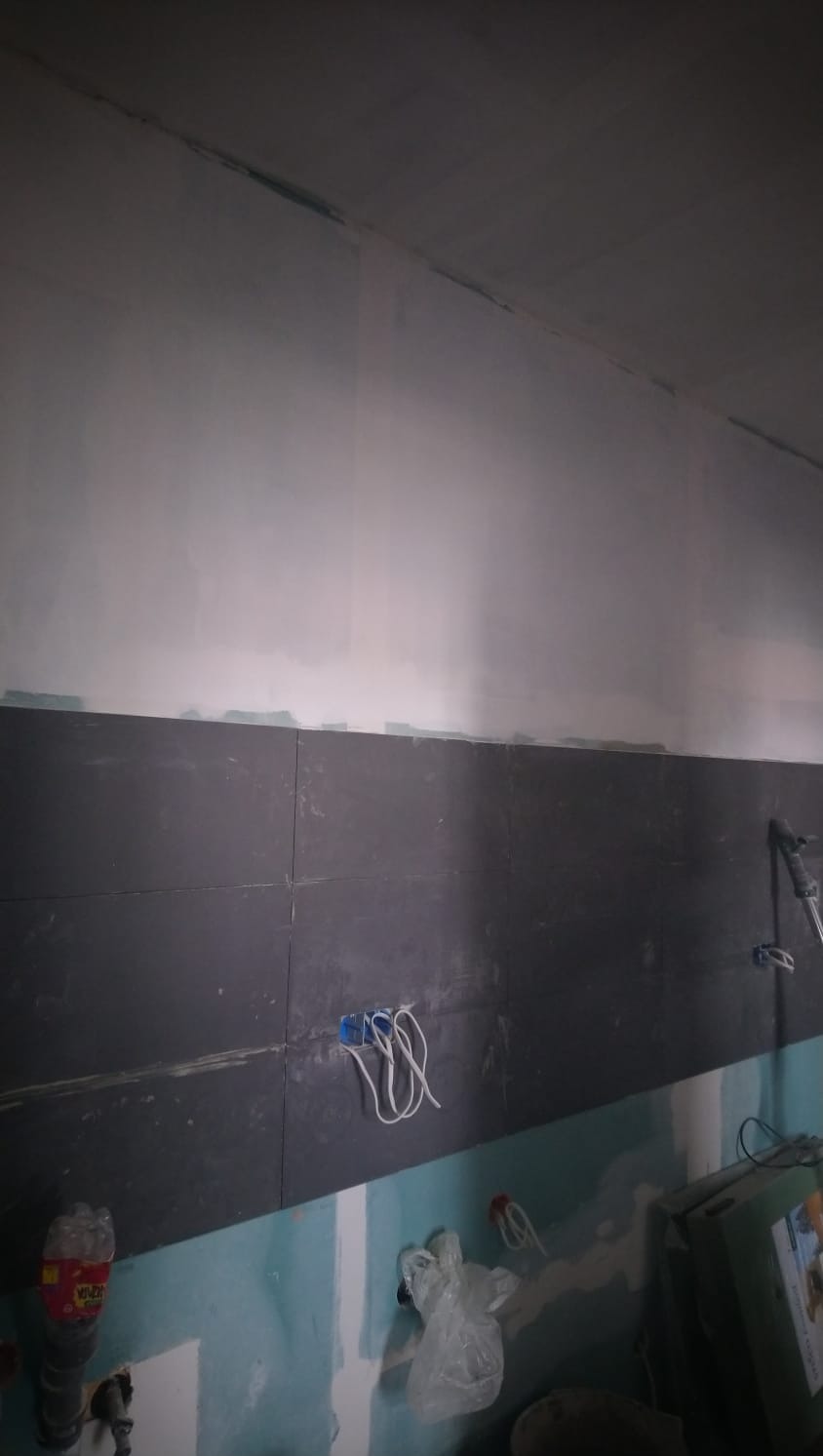 Ściana w trakcie remontu: czarne płytki ułożone do połowy wysokości, górna część ściany zagruntowana, widoczne wystające przewody elektryczne z niebieskiej puszki, folia ochronna i butelka po...