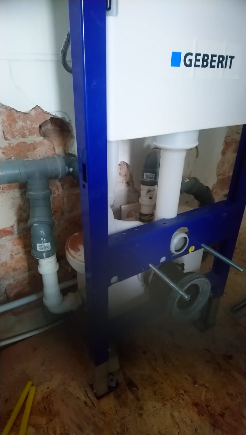 Stelaż podtynkowy Geberit zamontowany przy ścianie z fragmentami odsłoniętej cegły, widoczne rury kanalizacyjne i elementy mocujące, podłoga z płyty OSB.