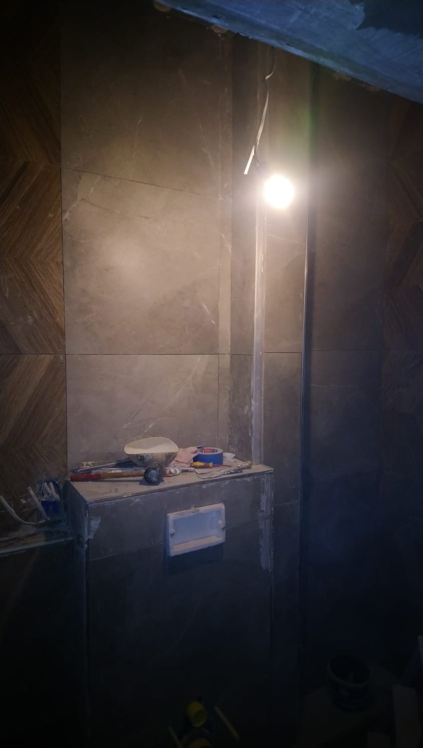 Wnętrze łazienki w trakcie prac wykończeniowych: płytki imitujące marmur na ścianach, narzędzia i materiały budowlane na zabudowie stelaża podtynkowego WC, widoczne przewody elektryczne.