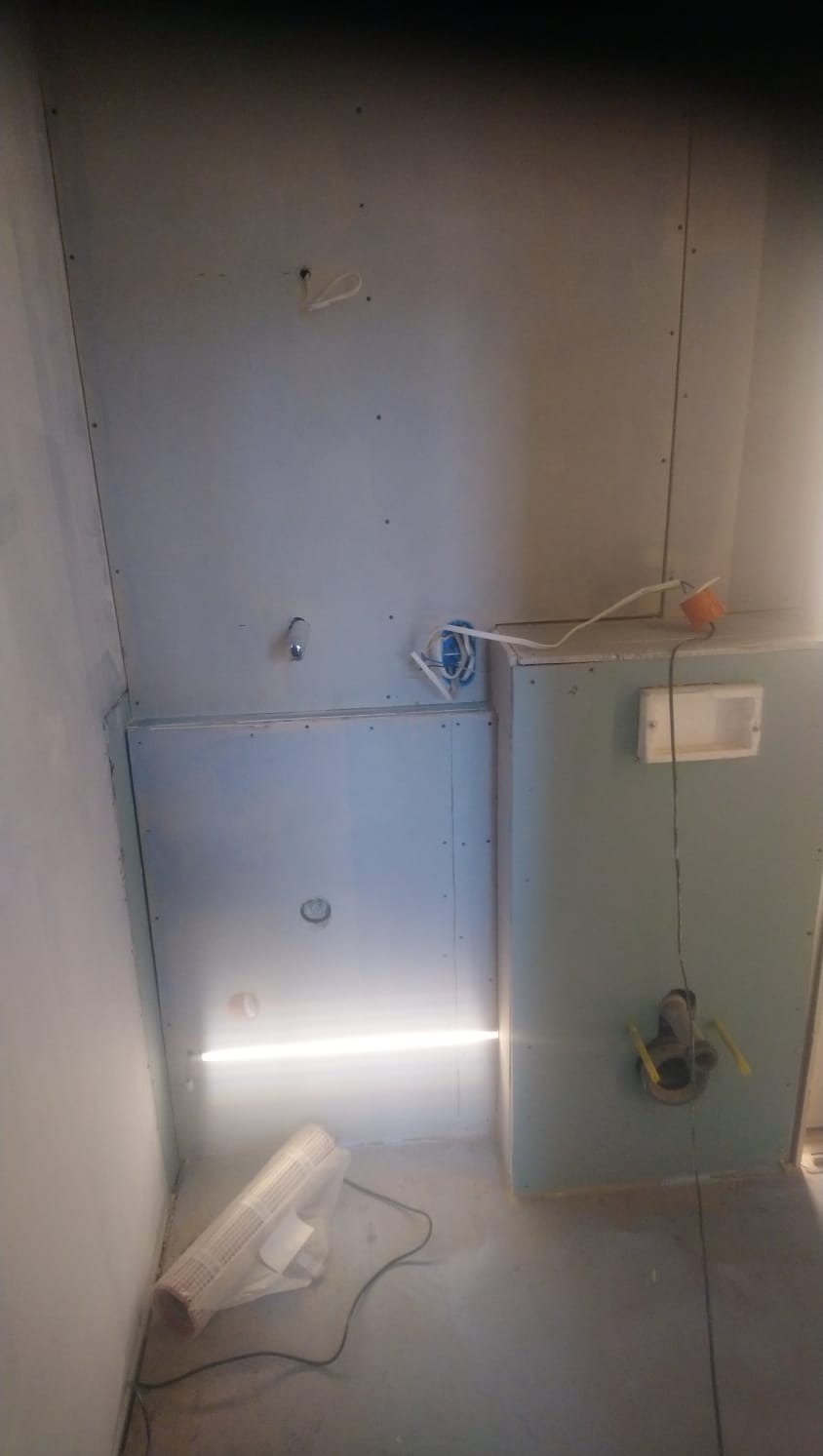 Wnętrze łazienki w trakcie prac wykończeniowych: zamontowane płyty gipsowo-kartonowe, widoczne puszki elektryczne, instalacja wodna, przewody oraz rura kanalizacyjna. Na podłodze leży zwinięta mata...