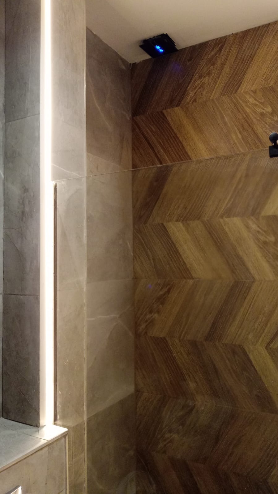 Fragment łazienki z drewnianą ścianą ułożoną we wzór jodełki, szarą płytką i pionowym oświetleniem LED. Widoczny czujnik z niebieskimi diodami na suficie.