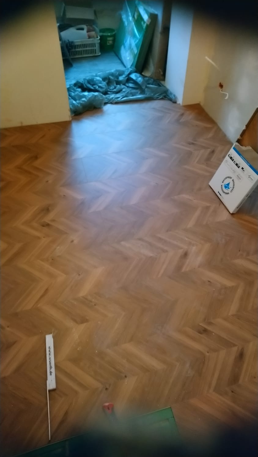 Ułożona drewniana podłoga w jodełkę, widoczne narzędzia pomiarowe i karton, w tle zabezpieczone folią elementy