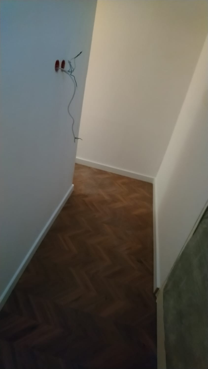 Wnętrze po odnowieniu: parkiet ułożony w jodełkę, świeżo pomalowane białe ściany z widocznymi przewodami elektrycznymi wychodzącymi ze ściany, białe listwy przypodłogowe.