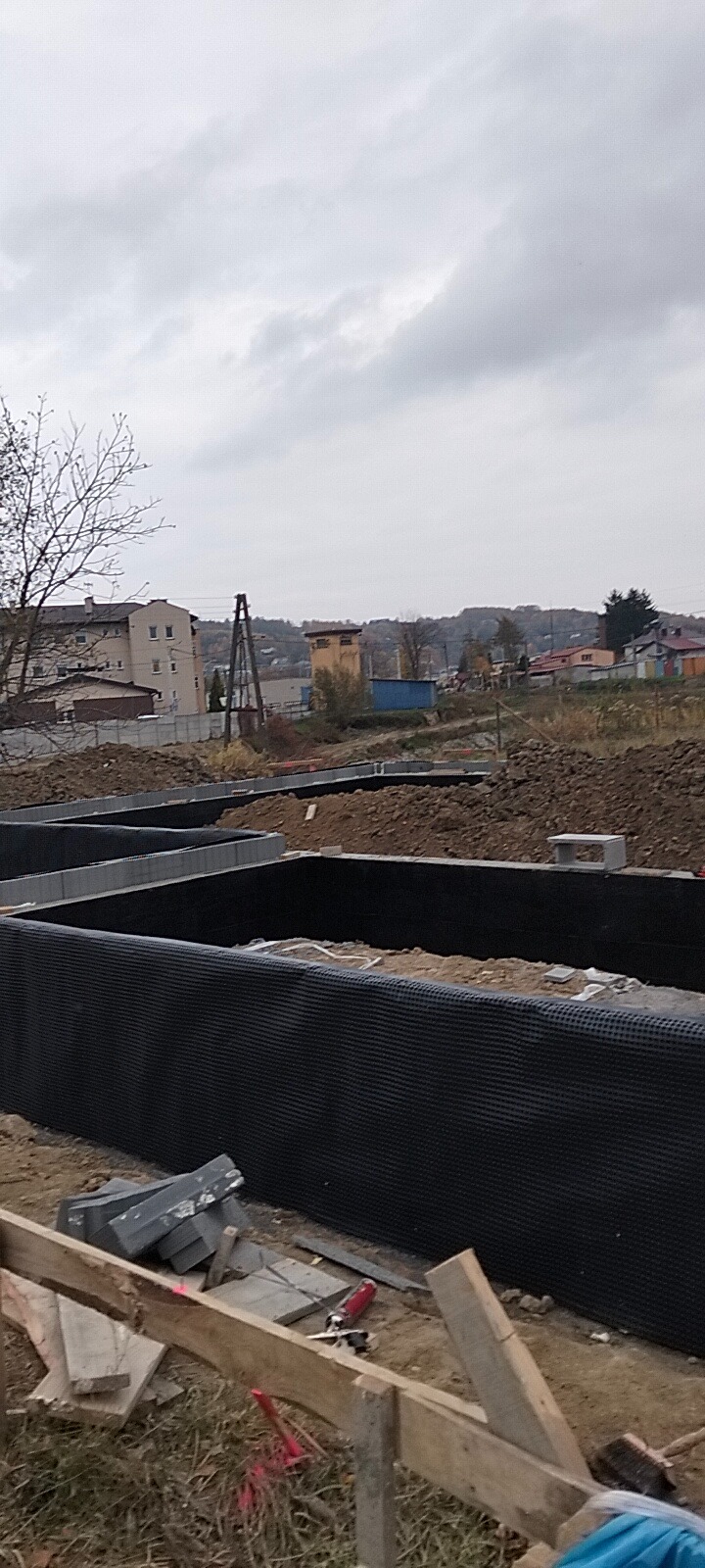 Zabezpieczone folią fundamenty domu w trakcie budowy, widoczne szalunki i materiały budowlane, w tle zabudowa mieszkalna i pochmurne niebo.