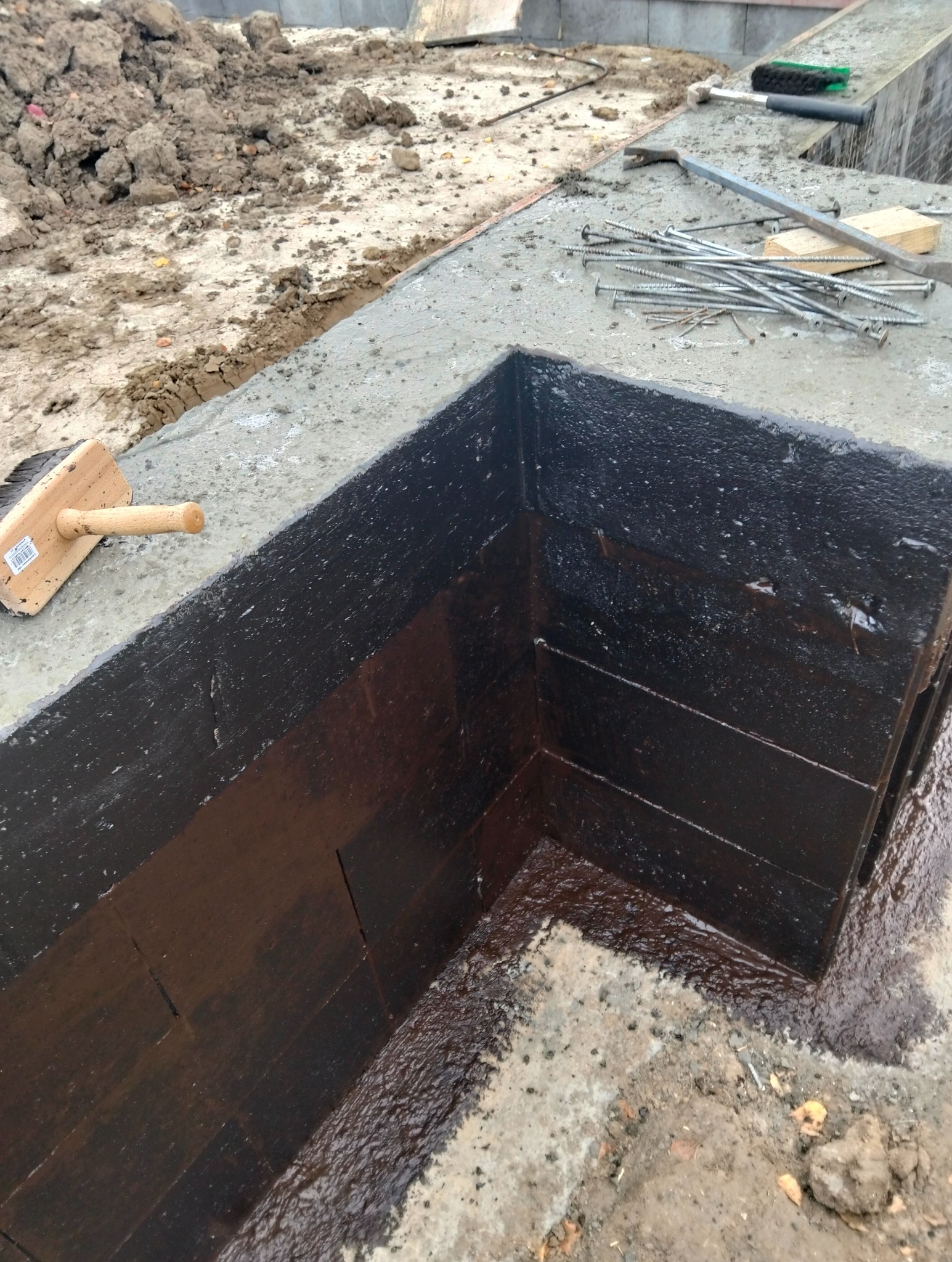 Hydroizolacja pionowa fundamentu z widoczną warstwą bitumiczną, pędzlem dekarskim i rozsypanymi gwoździami na betonie