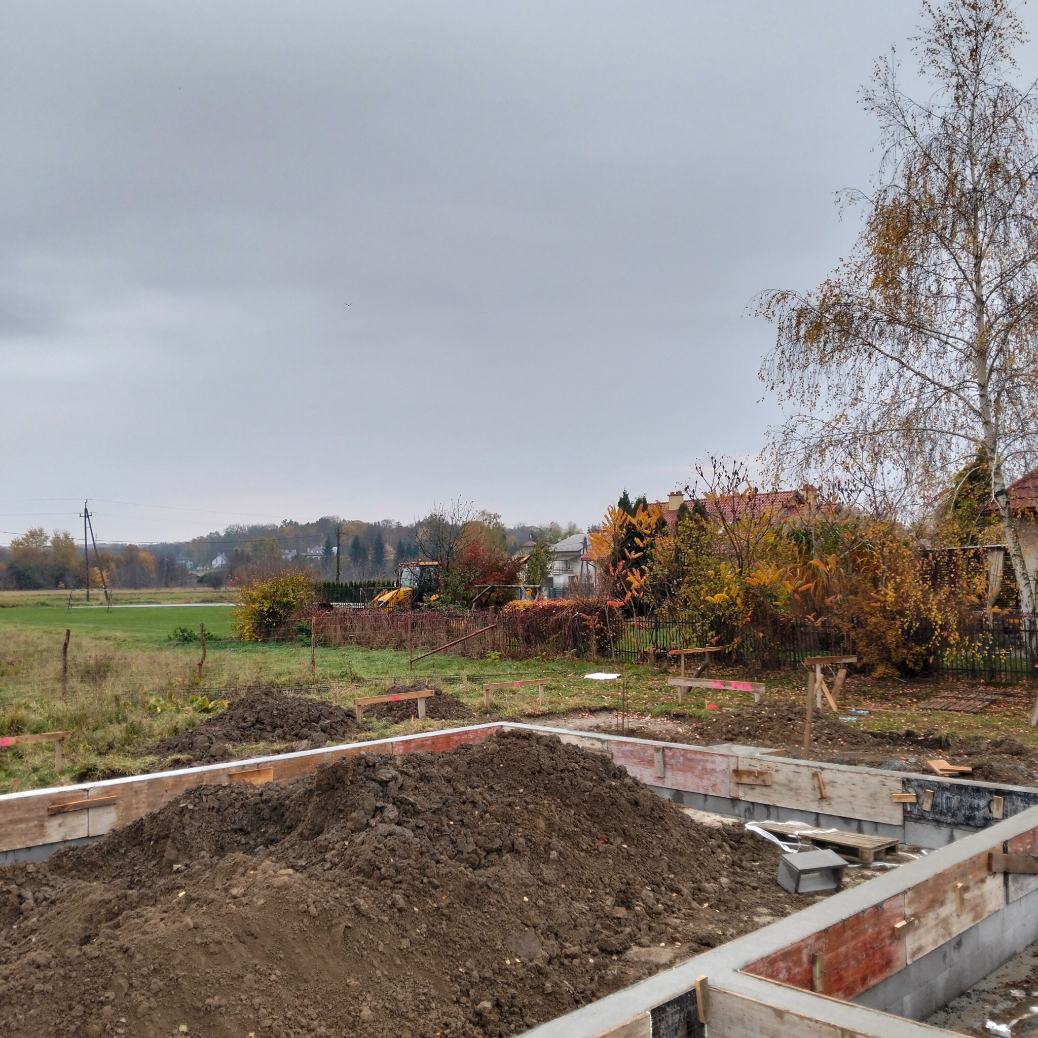 Zalane betonem fundamenty z drewnianym szalunkiem na tle jesiennego krajobrazu i koparki.