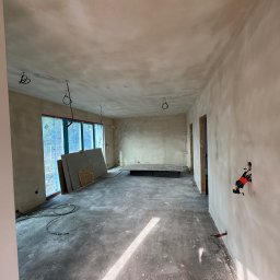 PaintCraft - Wnętrze w trakcie prac wykończeniowych: ściany pokryte gładzią, widoczne przewody elektryczne, płyty karton-gips opierające się o okno, surowa podłoga z plamami. Na ścianie zamontowane puszki...