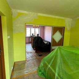 PaintCraft - Przygotowania do odświeżenia ścian w Białymstoku: pomieszczenie z częściowo zagruntowanymi, jaskrawozielonymi ścianami, podłoga zabezpieczona folią malarską, meble przykryte folią ochronną...