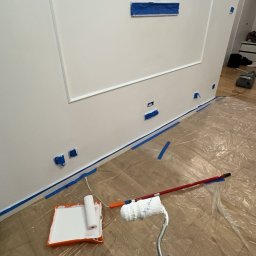 PaintCraft - Przygotowanie do malowania ściany: biała ściana z dekoracyjną ramą zabezpieczona taśmą malarską, podłoga zabezpieczona folią, wałek do malowania w dłoni, kuweta z farbą obok.