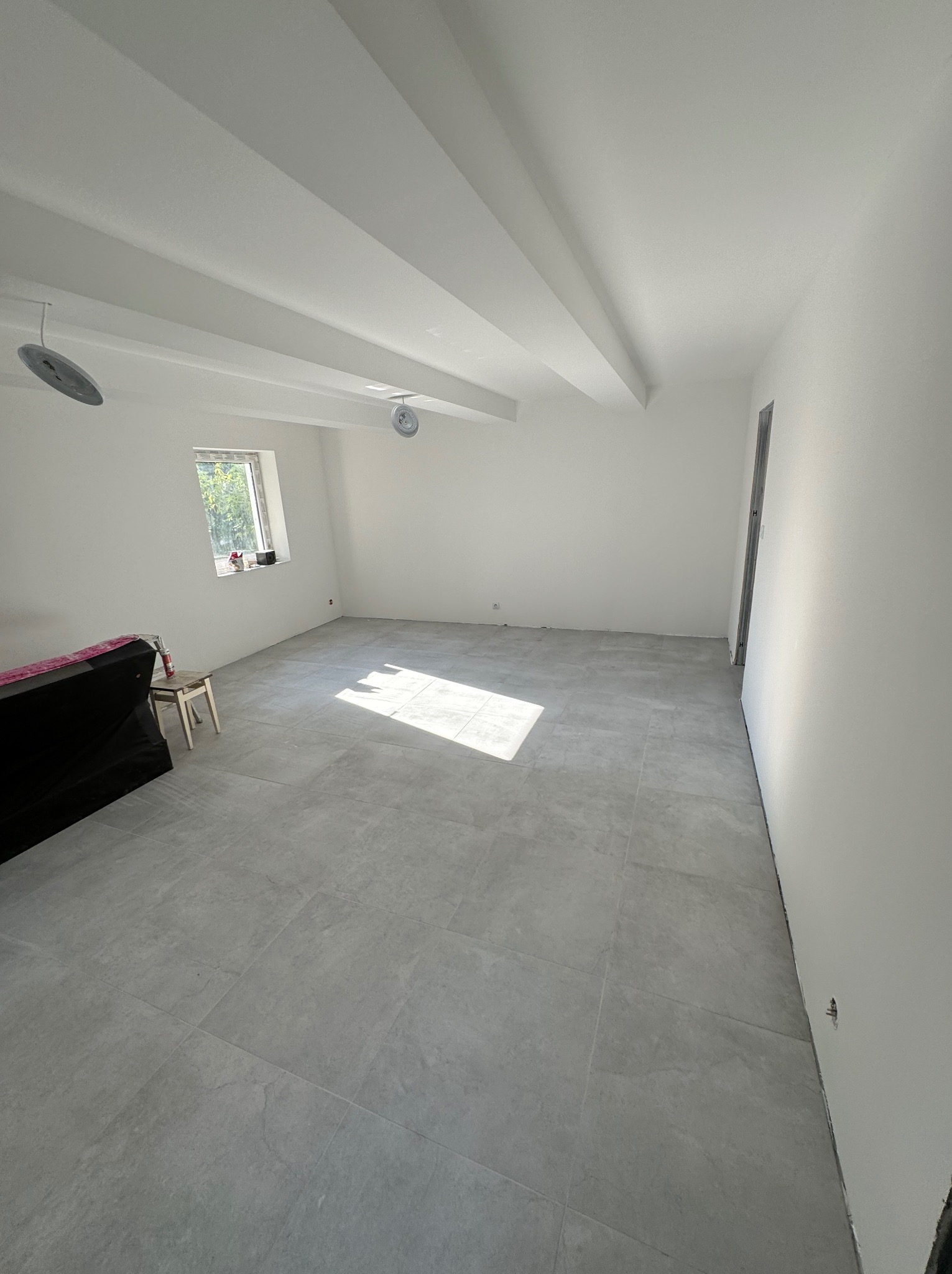 Jasne, przestronne wnętrze po remoncie z szarymi płytkami podłogowymi, białe ściany i sufit z widocznymi belkami oraz lampami, okno z widokiem na zieleń na zewnątrz, meble częściowo zakryte folią...