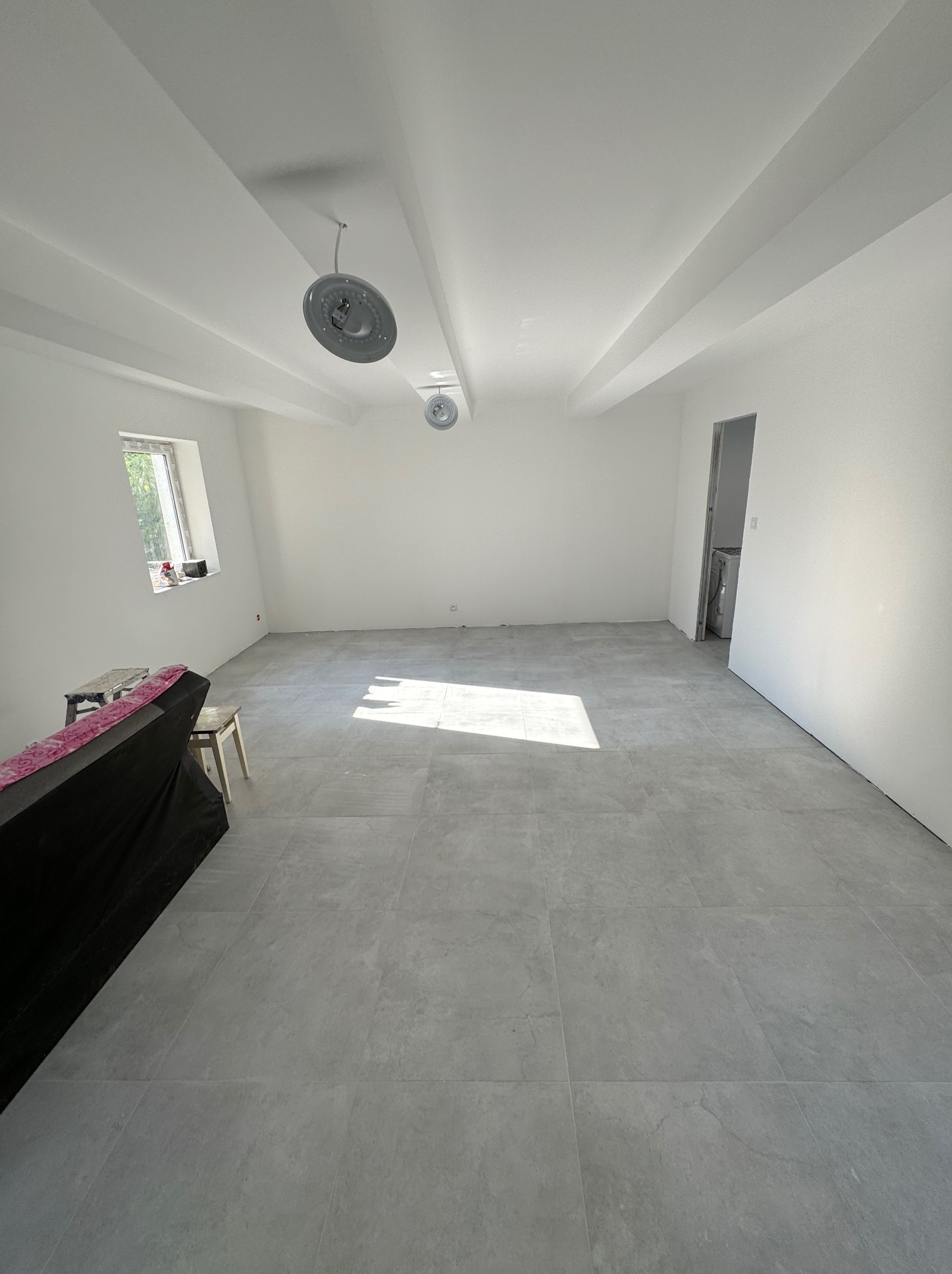 Wnętrze po remoncie: szare płytki podłogowe, świeżo pomalowane białe ściany i sufit z widocznymi belkami konstrukcyjnymi, niedokończone podłączenie lamp sufitowych.