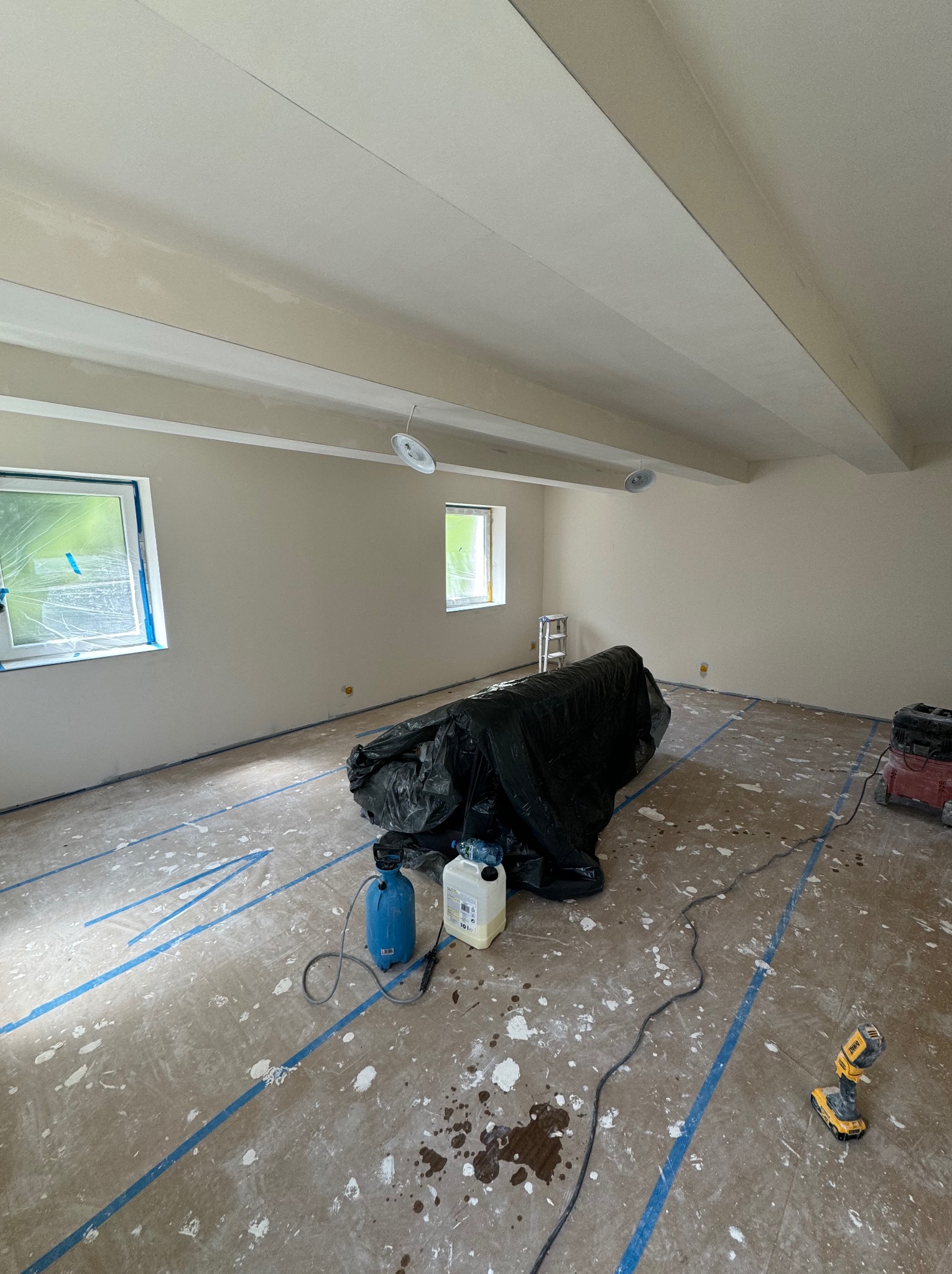 Przygotowane pomieszczenie do prac wykończeniowych: zabezpieczona podłoga taśmą malarską i folią, ściany w jasnym odcieniu, widoczne narzędzia i sprzęt malarski.