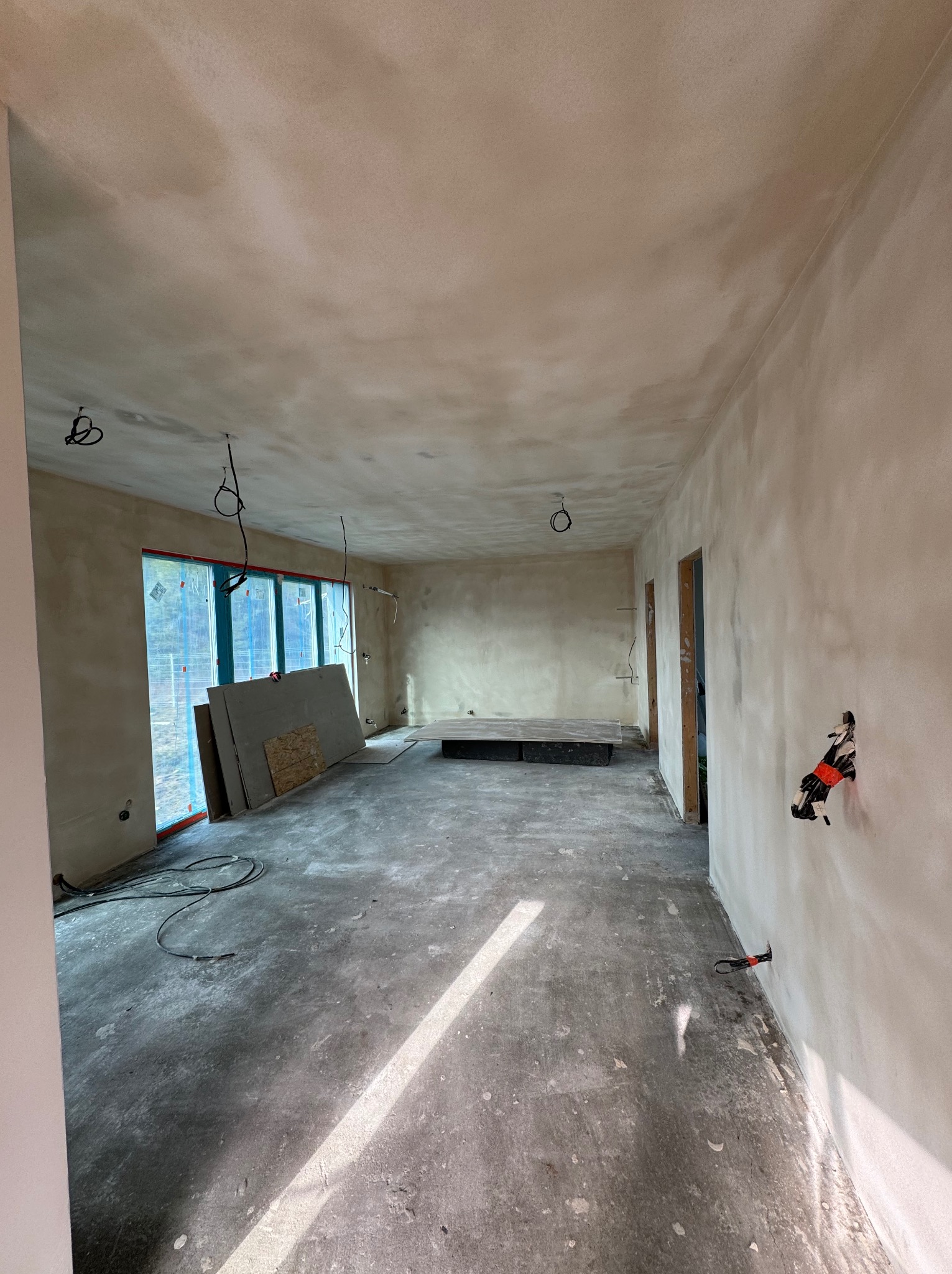 Wnętrze w trakcie prac wykończeniowych: ściany pokryte gładzią, widoczne przewody elektryczne, płyty karton-gips opierające się o okno, surowa podłoga z plamami. Na ścianie zamontowane puszki...