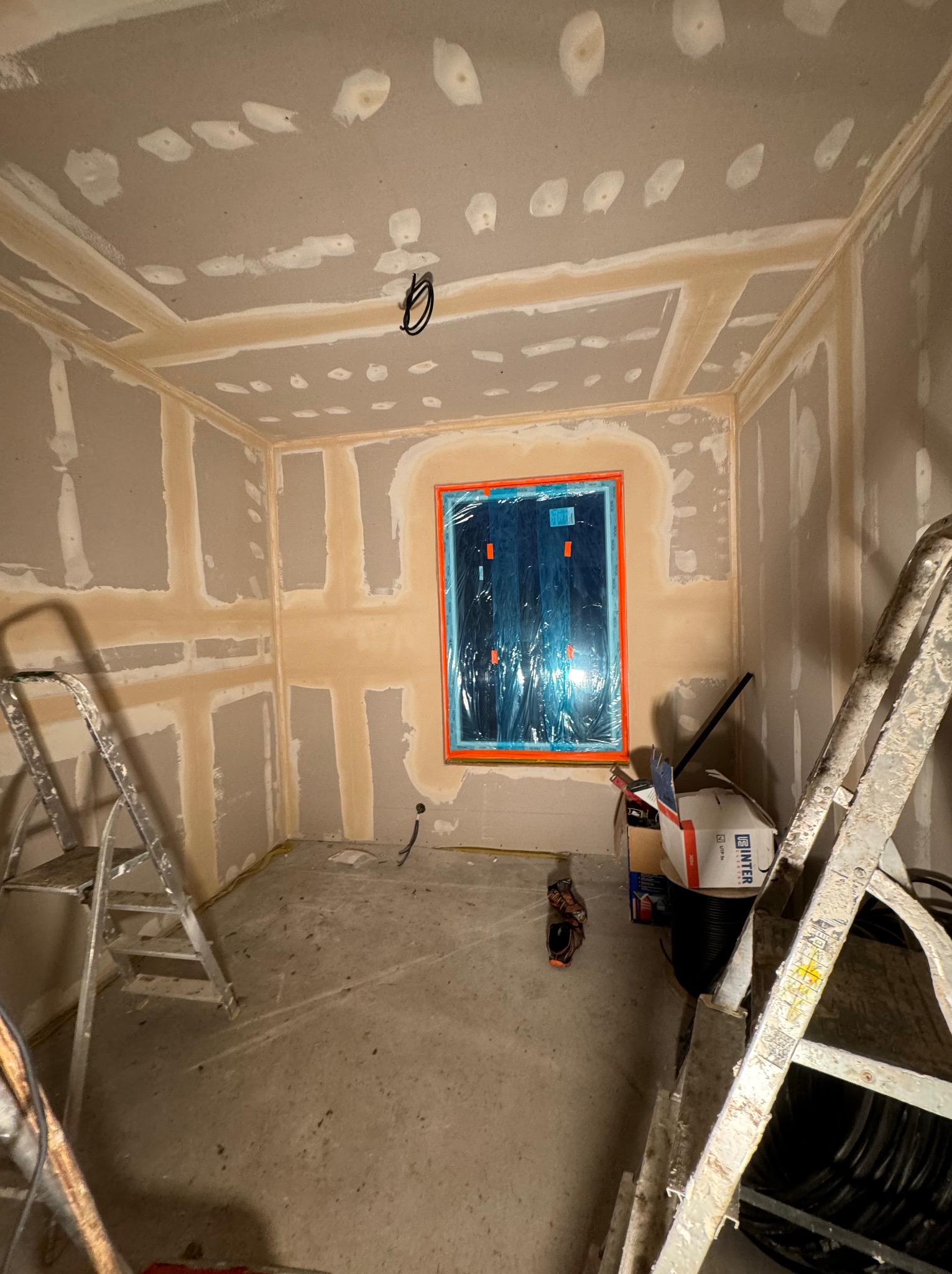 Pokój w trakcie prac przygotowawczych do wykończenia ścian, widoczne płyty kartonowo-gipsowe na ścianach i suficie, zaszpachlowane łączenia, okno zabezpieczone folią, drabiny w pomieszczeniu.