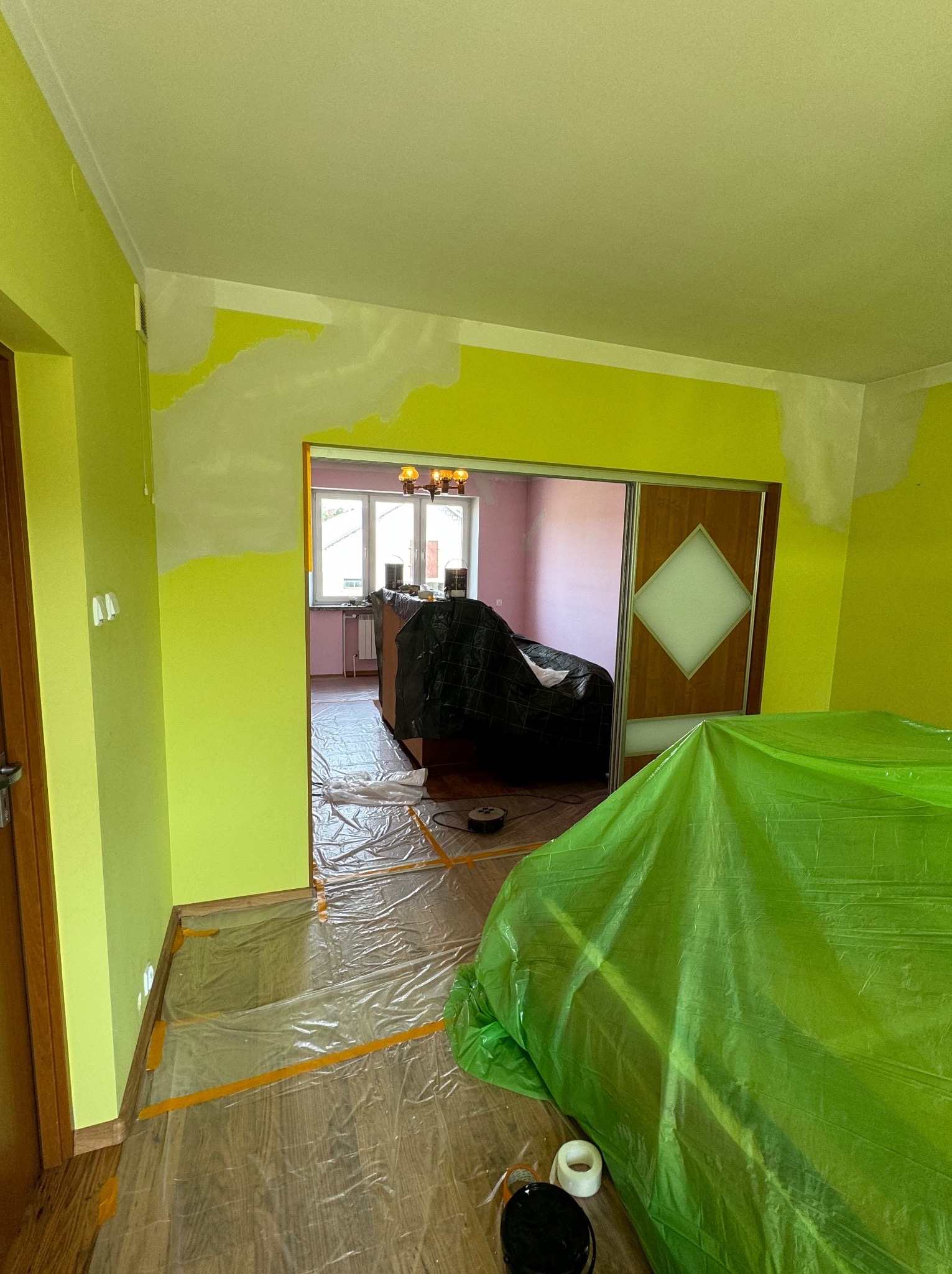 Przygotowania do odświeżenia ścian w Białymstoku: pomieszczenie z częściowo zagruntowanymi, jaskrawozielonymi ścianami, podłoga zabezpieczona folią malarską, meble przykryte folią ochronną...