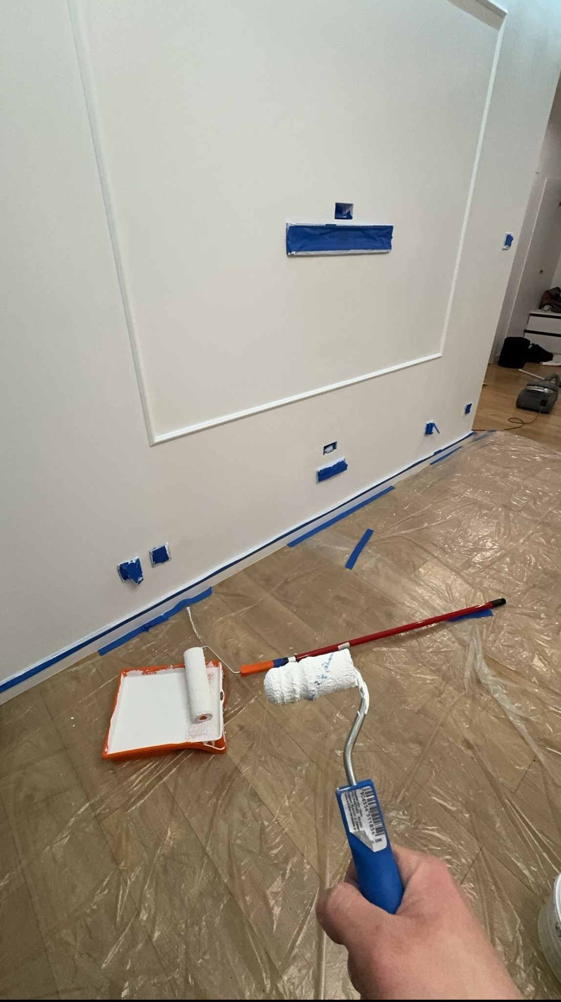 Przygotowanie do malowania ściany: biała ściana z dekoracyjną ramą zabezpieczona taśmą malarską, podłoga zabezpieczona folią, wałek do malowania w dłoni, kuweta z farbą obok.