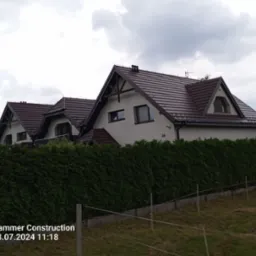 Dwa domy jednorodzinne z brązowymi dachami z dachówki ceramicznej, widoczne zza wysokiego żywopłotu, pochmurne niebo w tle.