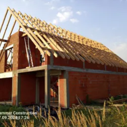 Surowy budynek z czerwonej cegły z drewnianą konstrukcją dachu w trakcie budowy, drabina oparta o ścianę, wysoka trawa na pierwszym planie, błękitne niebo w tle.