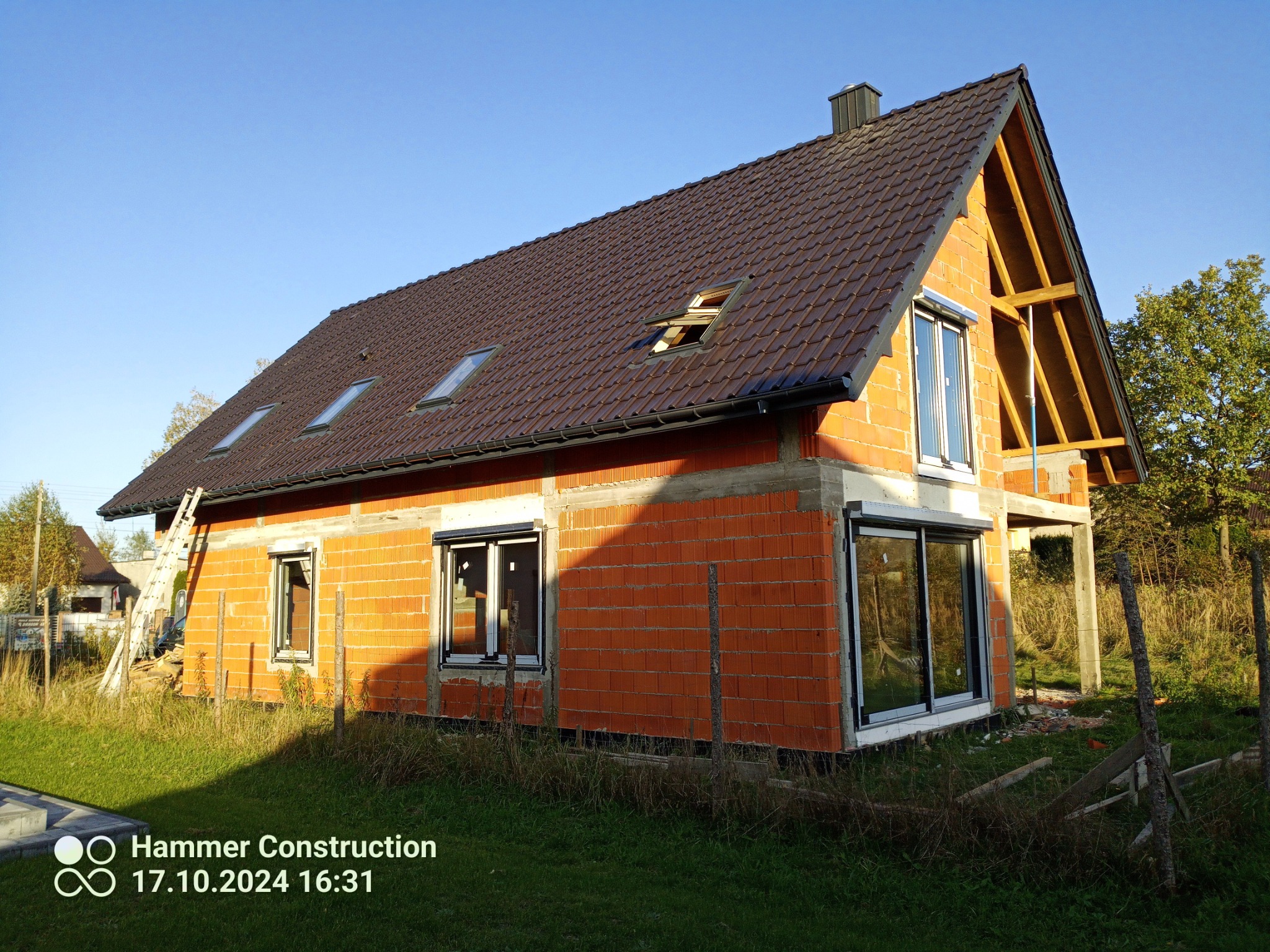 Dom w trakcie budowy z częściowo ułożonym brązowym pokryciem dachowym, widocznymi oknami dachowymi i elementami drewnianej konstrukcji szczytowej