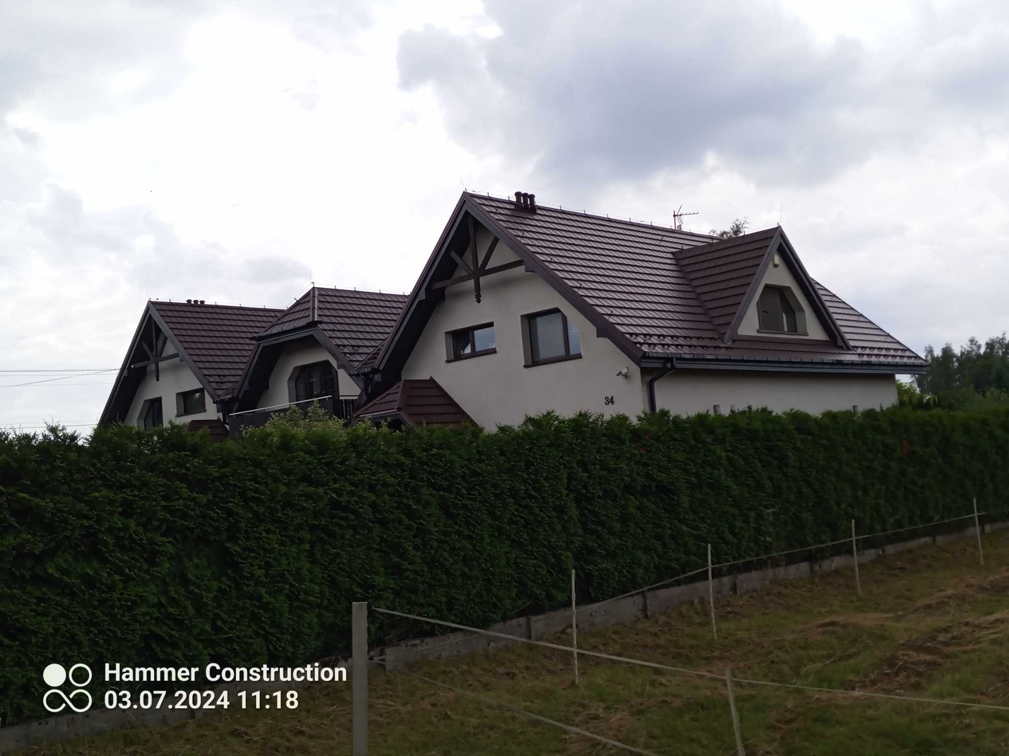 Dwa domy jednorodzinne z brązowymi dachami z dachówki ceramicznej, widoczne zza wysokiego żywopłotu, pochmurne niebo w tle.