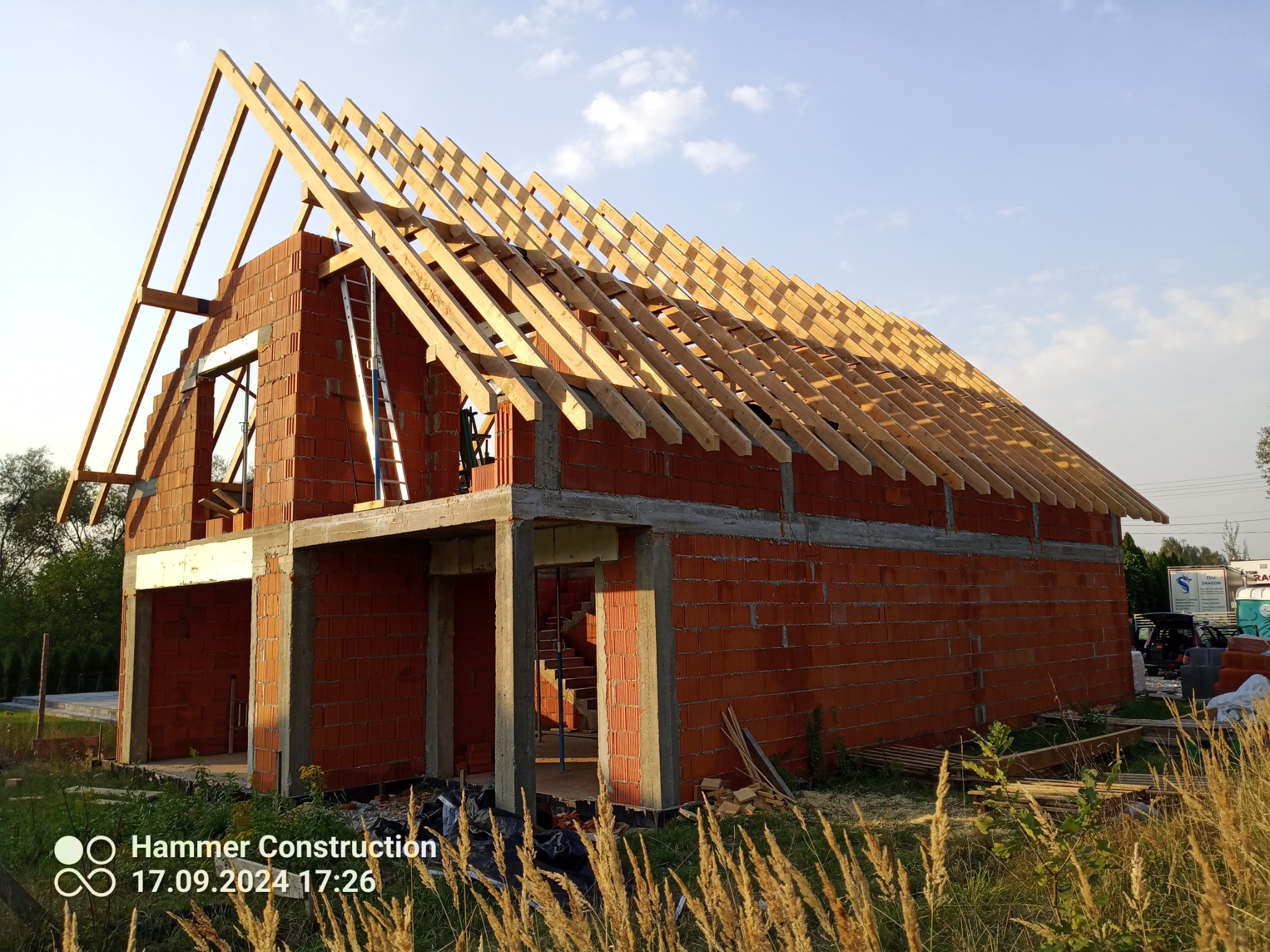 Surowy budynek z czerwonej cegły z drewnianą konstrukcją dachu w trakcie budowy, drabina oparta o ścianę, wysoka trawa na pierwszym planie, błękitne niebo w tle.