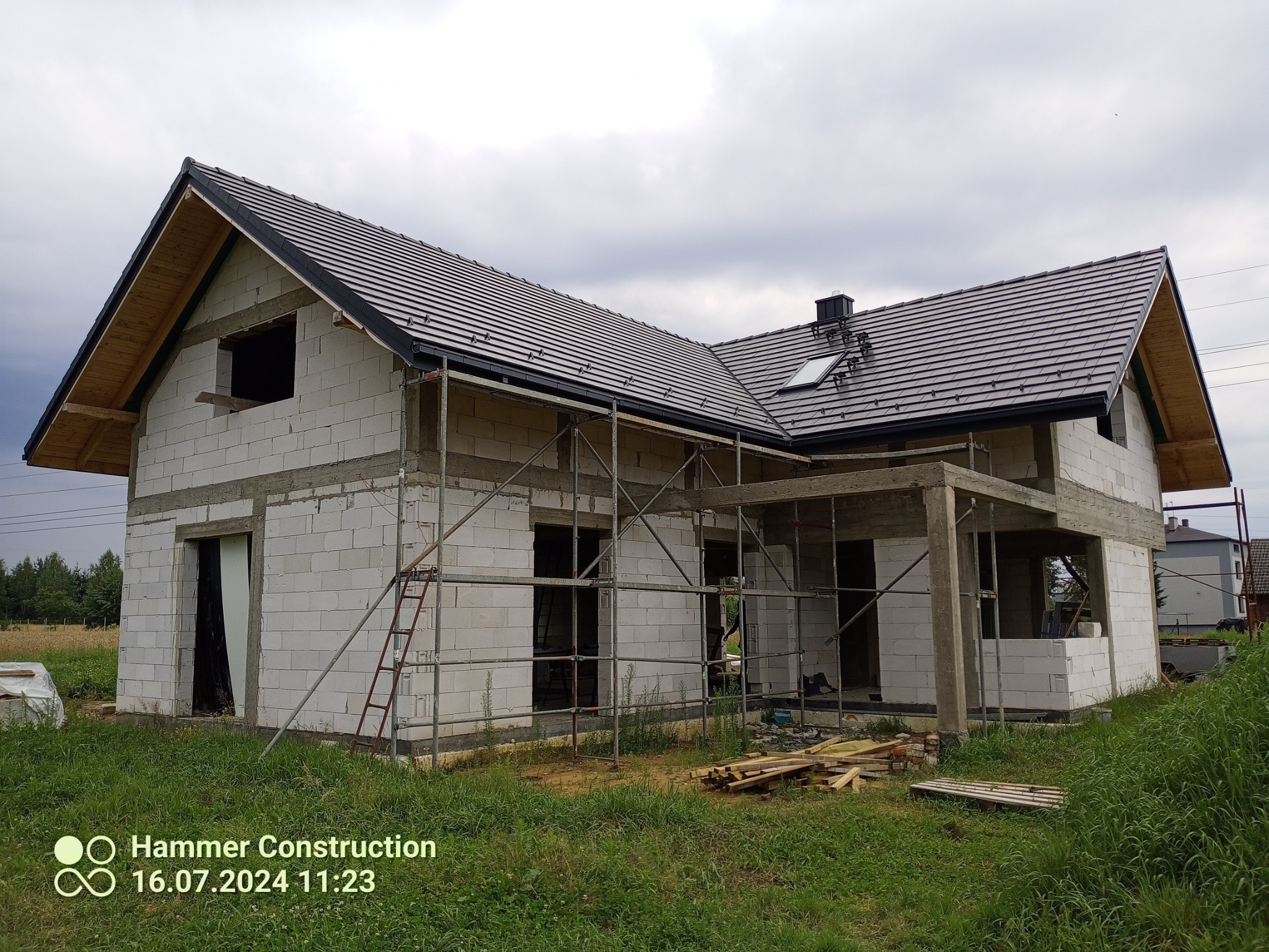 Dom w trakcie budowy z częściowo położonym, ciemnoszarym dachem z dachówki ceramicznej, widoczne rusztowania i materiały budowlane na trawniku.