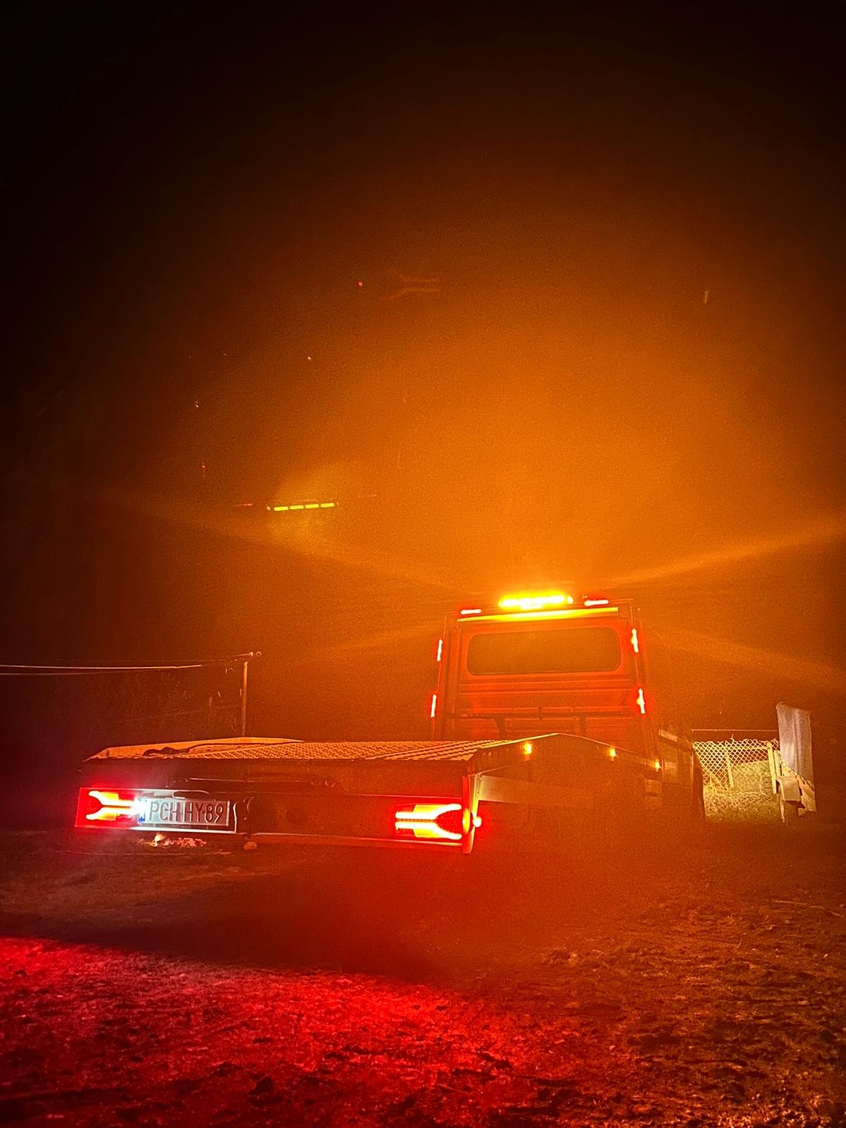 Ciężarówka z platformą transportową widziana z tyłu w nocy, podświetlona czerwonymi i pomarańczowymi światłami, tablica rejestracyjna widoczna.