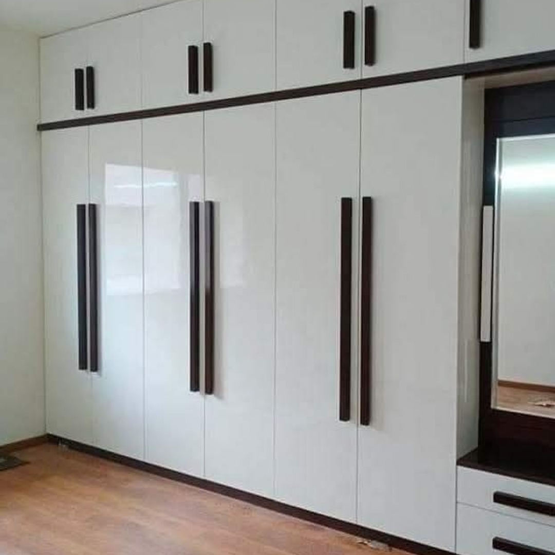 Biała szafa wnękowa z połyskującymi frontami i długimi, pionowymi uchwytami w kolorze ciemnego drewna, uzupełniona toaletką z lustrem po prawej stronie.