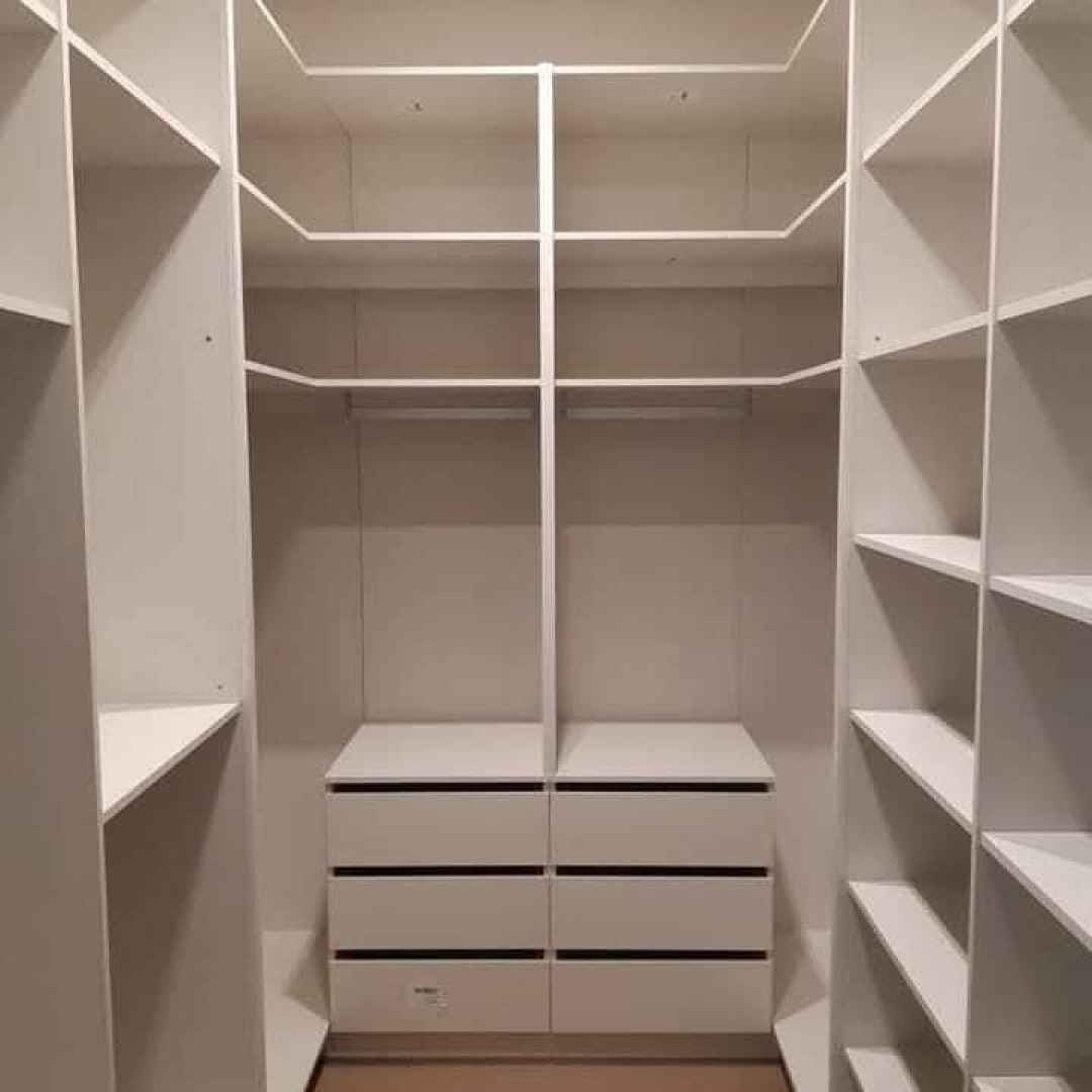 Wnętrze garderoby na wymiar z białymi szafkami i półkami, widoczne szuflady z czarnymi uchwytami, oświetlenie sufitowe.