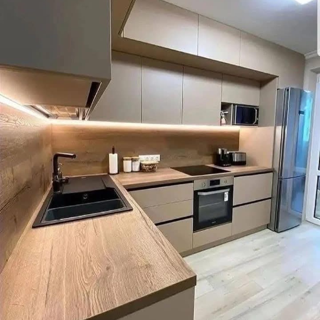 Nowoczesna kuchnia w odcieniach beżu i drewna, z czarnym zlewem i armaturą, oświetleniem LED pod szafkami wiszącymi i sprzętami AGD w zabudowie.