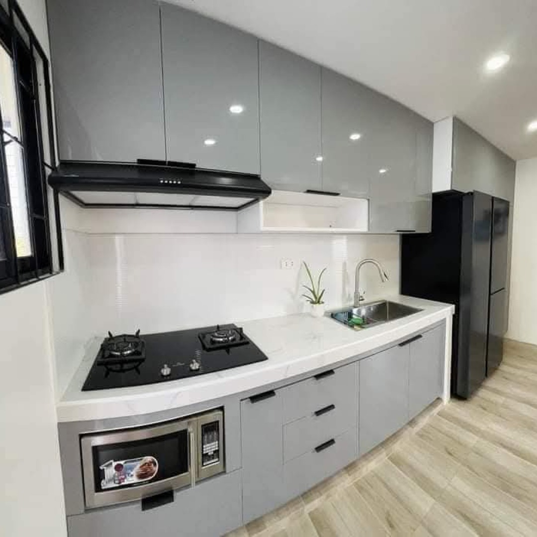 Nowoczesna, szara kuchnia z błyszczącymi frontami szafek, czarną płytą grzewczą i lodówką, oraz blatem imitującym marmur. W tle widoczne jasne panele podłogowe.