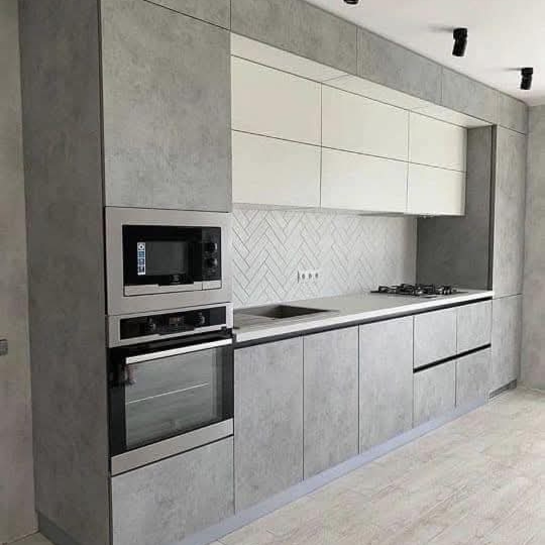 Nowoczesna kuchnia z szarymi, betonowymi frontami szafek i białymi szafkami wiszącymi, z mikrofalówką i piekarnikiem w zabudowie oraz białymi płytkami ułożonymi w jodełkę nad blatem.