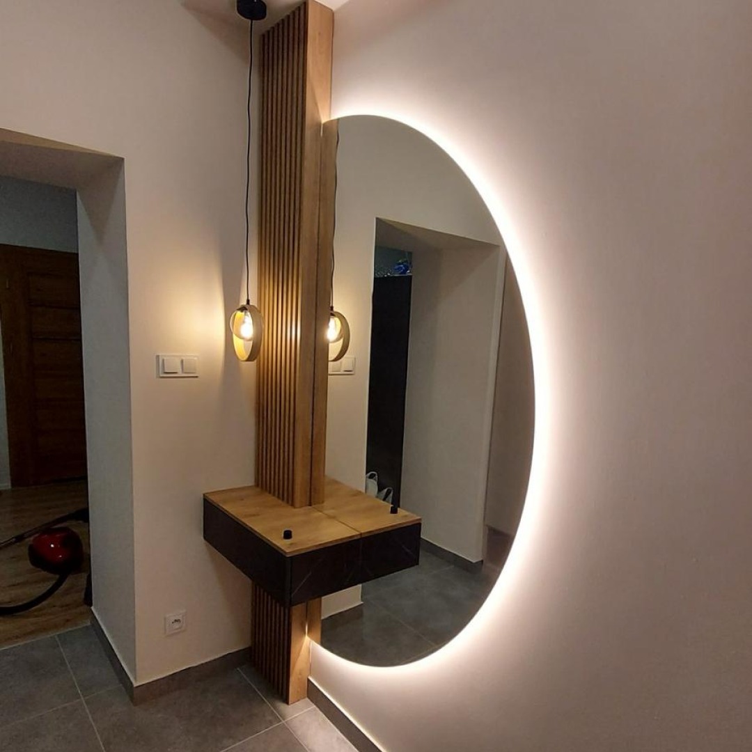 Nowoczesna aranżacja przedpokoju z okrągłym lustrem podświetlonym LED, drewnianą konsolą z szufladami i dekoracyjnym oświetleniem wiszącym.