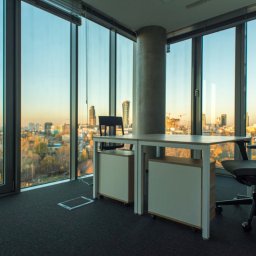 e-Support Sp. z o.o. - Nowoczesne biuro z widokiem na miasto o zachodzie słońca, minimalistyczne biurko z szafkami na kółkach i ergonomiczne krzesło biurowe, szara wykładzina dywanowa i betonowy filar.